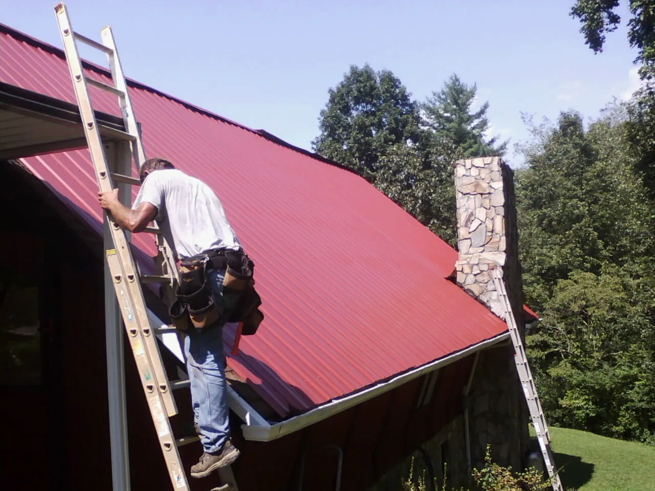 Roofer on a ladder beside a red metal roof and stone chimney — Soffit & Fascia Repair by Roofing Brothers in Denville